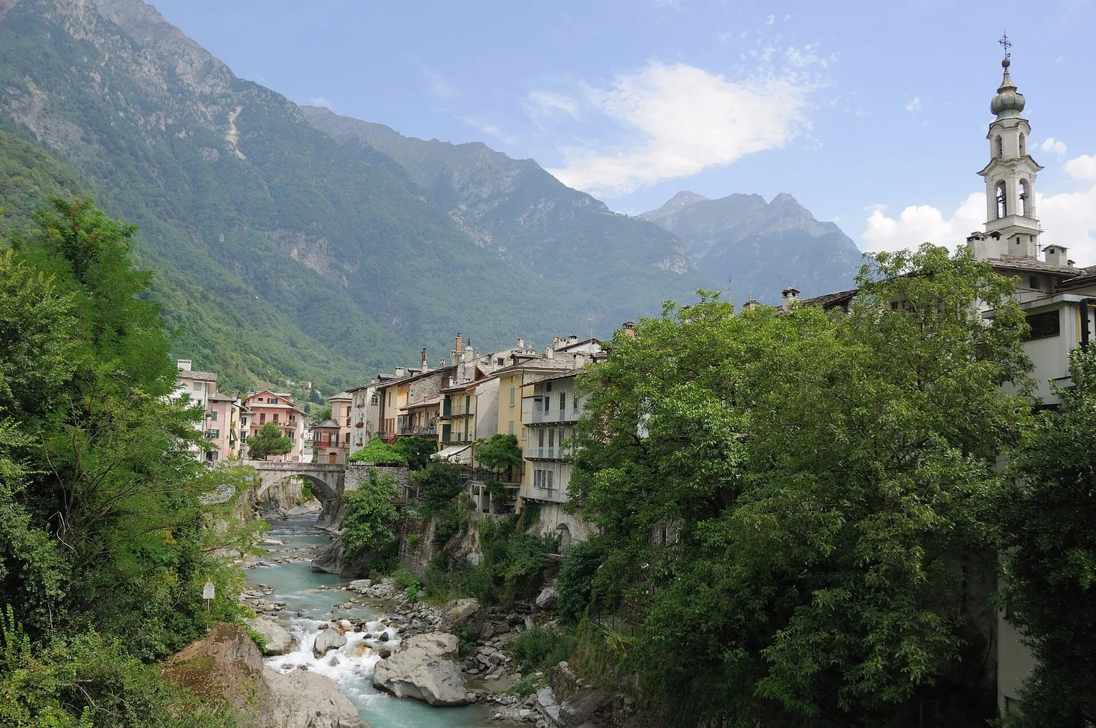 Il fiume Mera che scorre tra i massi nel centro di Chiavenna, con le case affacciate sull'acqua, un ponte in pietra, il campanile bianco a destra e le montagne della Valchiavenna sullo sfondo