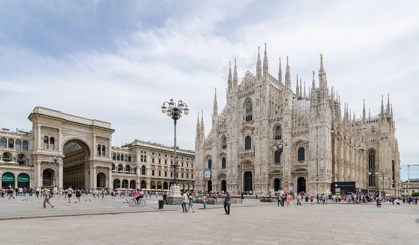 Piazza del Duomo a Milano: a destra la facciata gotica in marmo chiaro del Duomo con le guglie, a sinistra l'arco d'ingresso della Galleria Vittorio Emanuele II, al centro un grande lampione e passanti sulla piazza