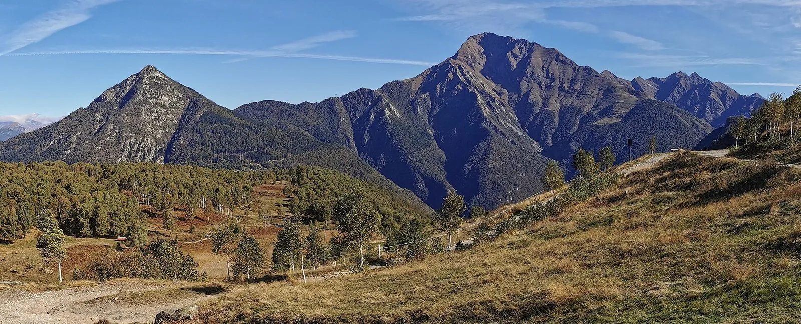 Panorama di montagna in autunno: a sinistra la piramide rocciosa del Legnoncino, a destra la mole più alta del Monte Legnone, davanti pascoli dorati con betulle e una strada sterrata