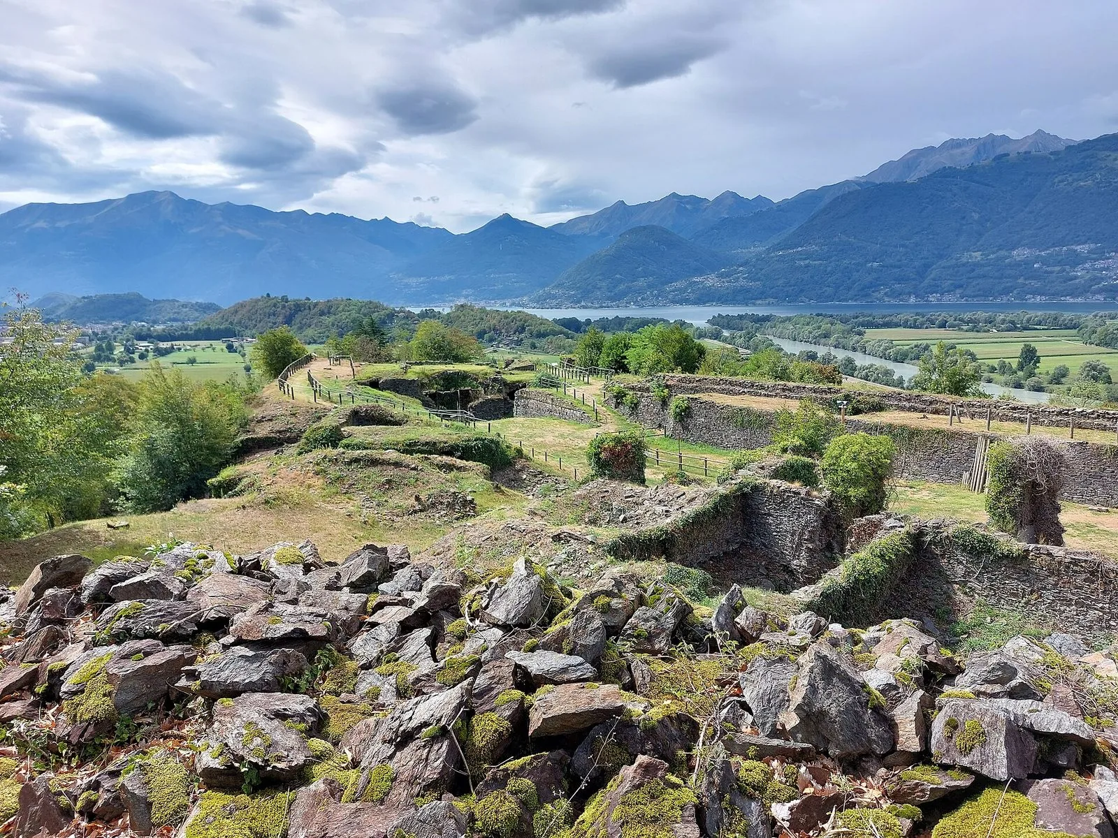 I ruderi in pietra del Forte di Fuentes coperti di muschio ed erba, con i camminamenti recintati; sullo sfondo la piana verde del Pian di Spagna, il corso del fiume, il lago e le montagne sotto un cielo nuvoloso
